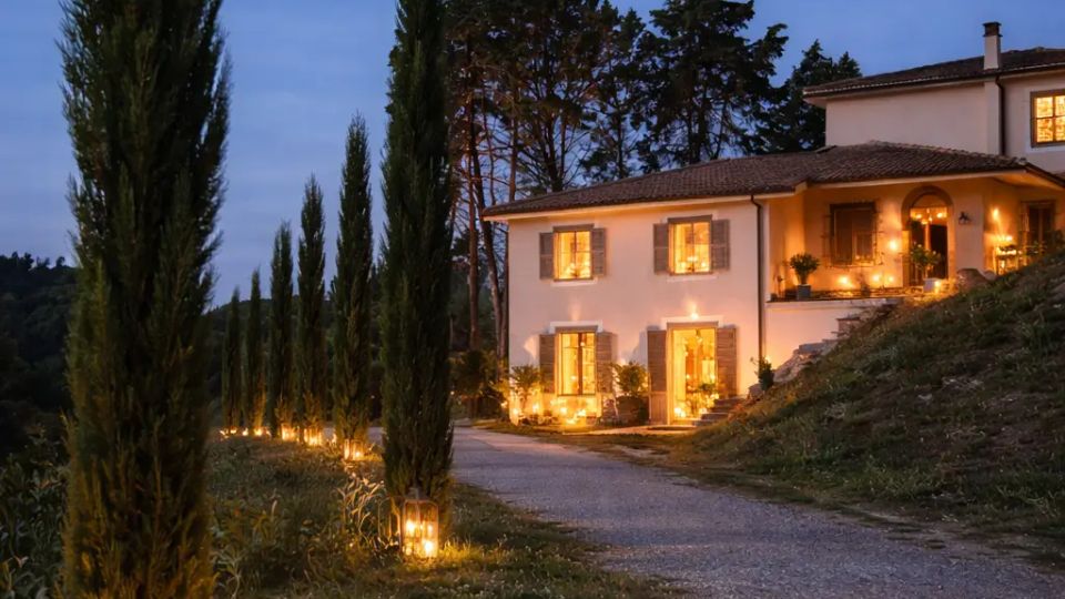 Viale di ghiaia in campagna fiancheggiato da alti cipressi, illuminato da lanterne a terra lungo il percorso. Sullo sfondo si vede una villa a due piani con persiane e luci calde accese, circondata da alberi sotto un cielo serale.