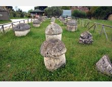 Area archeologica all’aperto con prato e una serie di blocchi in pietra a forma di cupola disposti in file, delimitata da staccionate in legno. Sullo sfondo si vedono un piccolo edificio e alberi che circondano il sito.