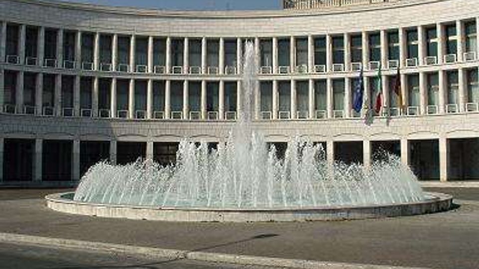 Ampia piazza con una grande fontana circolare a getti d’acqua al centro, davanti a un edificio moderno curvilineo in pietra chiara con portici e molte finestre. Sulla facciata sono visibili alcune bandiere vicino all’ingresso.