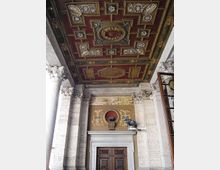 Soffitto a cassettoni riccamente decorato in rosso e oro sopra un portico in pietra con colonne e capitelli scolpiti. In basso si vede un ingresso con portale in pietra e doppia porta in legno, sormontato da un fregio ornamentale.