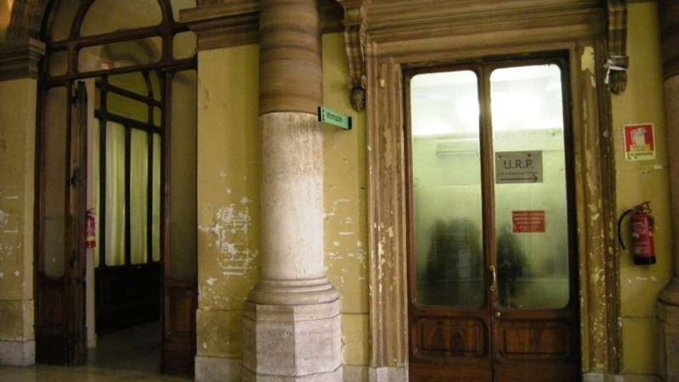 Ingresso interno di un edificio storico con pareti gialle consumate, colonne in pietra e cornici decorative attorno a porte in legno con vetri opachi. A sinistra c’è un grande arco vetrato che conduce a un corridoio, mentre a destra una porta con insegna “U.R.P.” è affiancata da segnaletica e un estintore a parete.