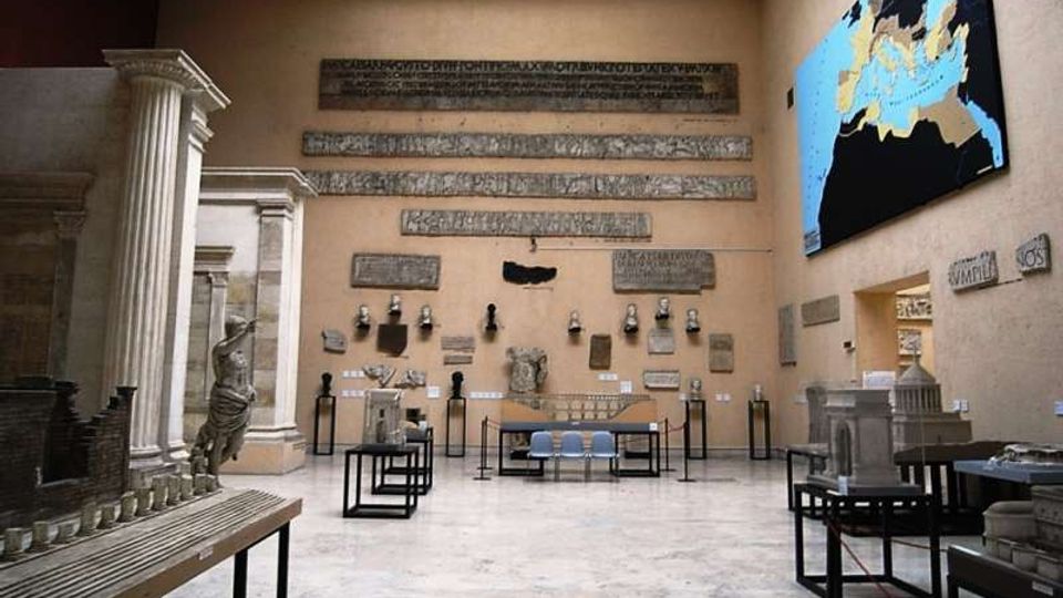 Sala museale con pareti beige e lucernario, con iscrizioni e frammenti lapidei antichi esposti a parete e su pedane. Ai lati si vedono modelli architettonici e reperti in vetrina, e sulla parete destra un grande pannello con una mappa.