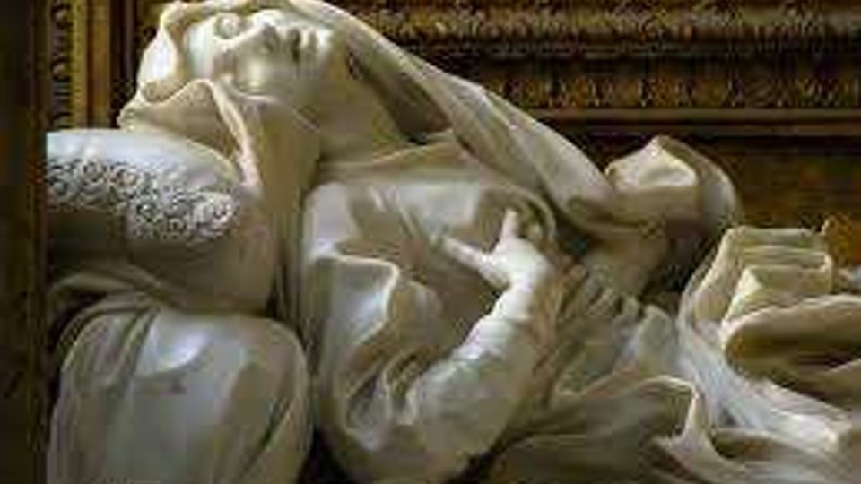 Scultura in marmo bianco raffigurante una figura distesa su un letto con drappeggi scolpiti e un velo sul volto, collocata in un ambiente interno con cornici decorative dorate sullo sfondo.