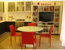 Interno open space con cucina moderna ad angolo, pensili con ante in vetro e piano di lavoro chiaro con forno e fornelli integrati. In primo piano c’è un tavolo rotondo con quattro sedie rosse, e sullo sfondo una grande libreria bianca con TV e scaffali pieni di libri e oggetti.