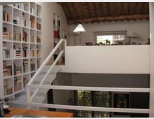 Interno di un loft su più livelli con soppalco e scala bianca, pareti chiare e grande libreria a tutta altezza piena di libri. Soffitto con travi in legno a vista, finestra sul fondo e affaccio su un livello inferiore con ampie superfici vetrate.