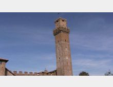 Torre medievale in pietra alta e slanciata, con merlature e coronamento sporgente in cima, ripresa contro un cielo azzurro. In basso si intravede una cinta muraria con merli e i tetti di edifici in muratura.