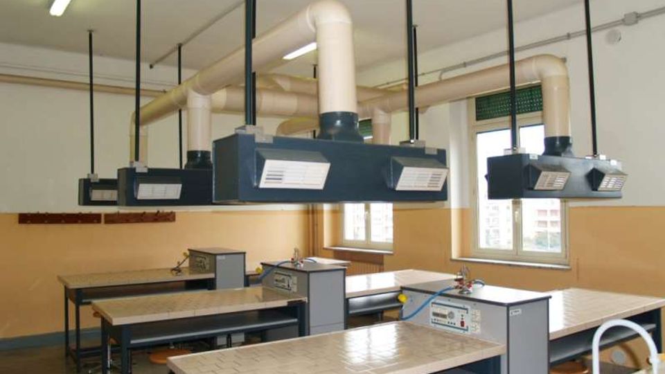Aula laboratorio con banchi da lavoro rivestiti in piastrelle e sgabelli, dotata di cappe aspiranti sospese collegate a grandi condotti di ventilazione. Sulle pareti chiare ci sono finestre e illuminazione a soffitto, con impianti e tubazioni visibili.