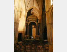 Interno di una chiesa in pietra con alte arcate gotiche e volte a crociera, illuminata da luce calda. In fondo si vede una cappella con baldacchino a cupola sopra un altare, con file di sedie in legno in primo piano.