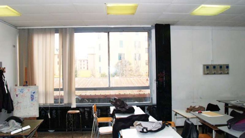 Aula scolastica con banchi e sedie disposti in modo irregolare, soffitto a pannelli con luci fluorescenti e pareti bianche. Sul fondo una grande finestra con tenda parziale affaccia su edifici esterni; presenti uno sgabello e alcuni pannelli/quadri appoggiati ai lati.