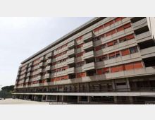 Grande edificio residenziale a più piani in cemento, con lunghe file di finestre e balconi e pannelli frangisole rossi. La struttura è sollevata su pilastri con un livello inferiore porticato e una passerella lungo la facciata.