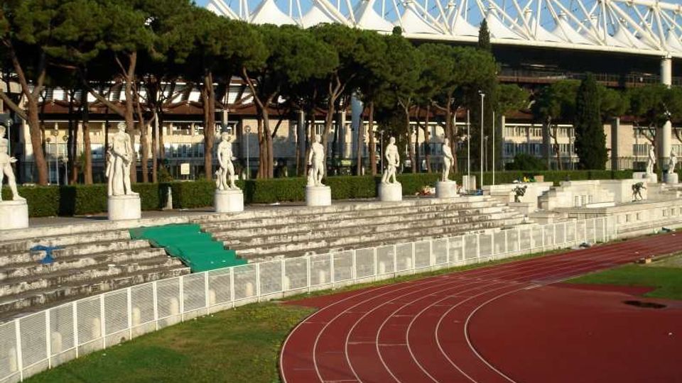 Stadio con pista di atletica rossa in curva e gradinate in cemento, lungo le quali sono allineate statue su piedistalli. Sullo sfondo si vedono una fila di pini e la struttura bianca a traliccio di una tribuna o copertura dello stadio.