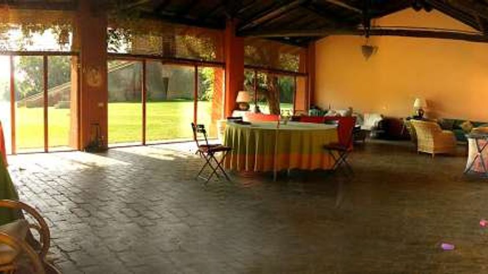 Ampio portico coperto con pavimento in pietra scura e travi in legno a vista, arredato con tavoli rotondi e sedute. Grandi vetrate e porte scorrevoli si affacciano su un prato verde e un’area esterna illuminata dal sole.