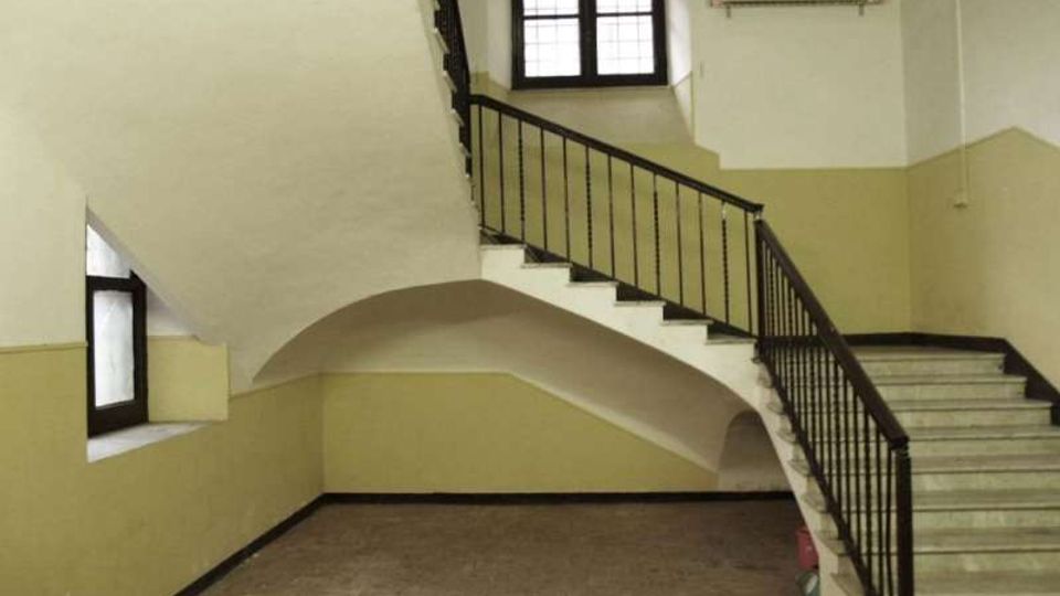 Interno di un vano scala in edificio, con pareti bicolore bianco e beige, pavimento in piastrelle e scalinata in pietra con ringhiera in ferro scuro. Una grande finestra quadrata e una piccola finestra laterale illuminano lo spazio, con un arco sotto la rampa principale.