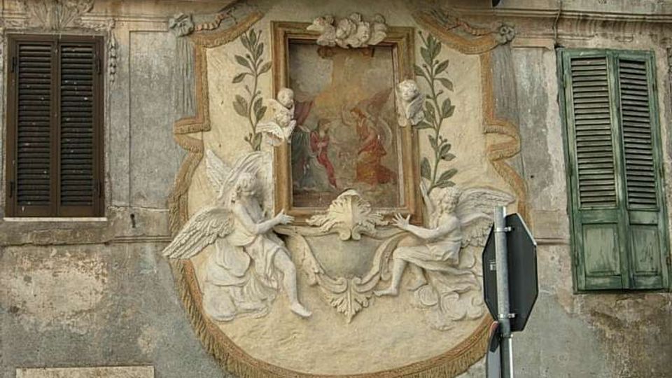 Facciata di un edificio storico con un riquadro decorativo in stucco che incornicia un piccolo affresco centrale, affiancato da figure angeliche e motivi vegetali in rilievo. Ai lati si vedono due finestre con persiane in legno e un cartello stradale in primo piano.
