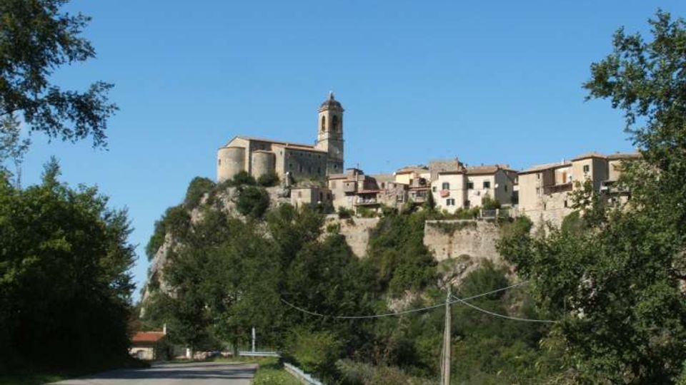 Borgo storico su uno sperone roccioso, con case in pietra e una chiesa con campanile al centro, circondato da alberi e mura di contenimento. In primo piano una strada asfaltata con guardrail e cavi elettrici sotto un cielo sereno.