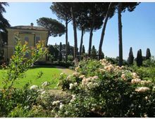 Giardino di una villa con prato curato e aiuole di rose in primo piano, affiancato da un edificio giallo con tetto in tegole. Sullo sfondo si vedono alti pini e cipressi che incorniciano il paesaggio sotto un cielo sereno.