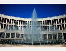 Fontana con alto getto d’acqua al centro di una piazza, davanti a un edificio moderno semicircolare con facciata a colonne e grandi finestre. Sullo sfondo si vede il cielo azzurro.