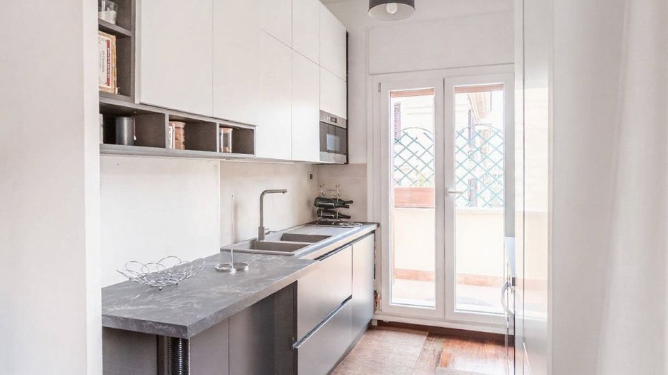 Cucina moderna e stretta in stile lineare, con mobili bianchi e grigi, piano di lavoro in pietra, lavello e piano cottura integrati e scaffali a vista. In fondo, una portafinestra a due ante illumina l’ambiente e si affaccia su un balcone con griglia decorativa.