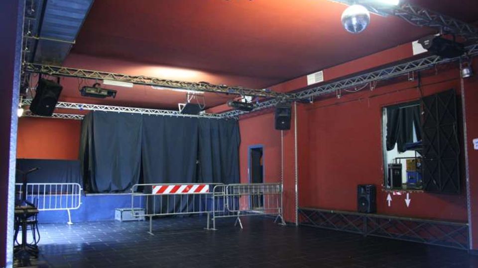 Interno di una piccola sala eventi con pareti rosso scuro e pavimento in piastrelle nere, con palco sul fondo chiuso da tende nere. Al soffitto ci sono tralicci con luci e altoparlanti, e davanti al palco sono presenti transenne metalliche.