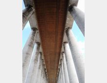 Vista dal basso di un viadotto in cemento armato, con l’intradosso marrone della carreggiata che corre in prospettiva tra alte colonne cilindriche. Il cielo azzurro è visibile ai lati, evidenziando la struttura ripetuta dei piloni.