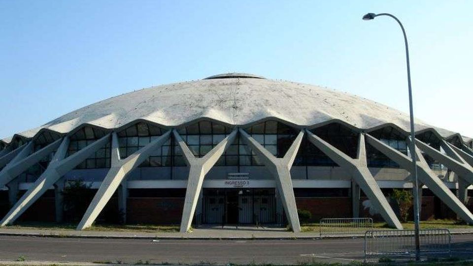 Vista frontale di un grande palazzetto dello sport con ampia copertura a cupola e strutture portanti esterne in cemento a forma di archi inclinati. Davanti si vede una strada con marciapiede e lampione, sotto un cielo sereno.
