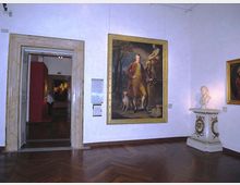 Sala di museo con pareti bianche e pavimento in parquet, con un grande dipinto incorniciato al centro e una scultura su piedistallo decorato sulla destra. A sinistra si vede un’ampia porta con cornice in pietra che conduce a un’altra galleria con pareti rosse e altre opere esposte.