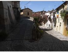Strada in pietra acciottolata in un borgo storico, con salite e curve tra case compatte in intonaco chiaro e balconi. Un’aiuola centrale con arbusti fioriti e alcuni tavolini all’aperto si affaccia su un piccolo slargo sotto un cielo sereno.