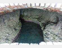 Apertura in muratura che affaccia su una cavità rocciosa con pareti irregolari e acqua limpida di colore turchese, simile a una piccola grotta o cisterna naturale. La vista è dall’alto, con i bordi in mattoni visibili lungo il perimetro.