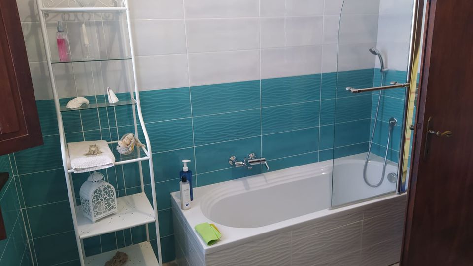 Bagno con vasca bianca dotata di parete in vetro e soffione doccia, rivestito da piastrelle bianche e una fascia turchese a effetto ondulato. Accanto alla vasca c’è una scaffalatura bianca con ripiani in vetro e accessori decorativi, e una porta in legno sul lato destro.