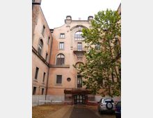 Cortile interno con edificio in mattoni a più piani, con finestre ad arco e un grande arco decorativo centrale sopra un balconcino. Un albero occupa il lato destro e l’ingresso al piano terra è coperto da una tettoia, con area pavimentata e auto parcheggiate.