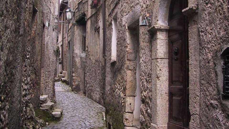 Stretto vicolo medievale in pietra con pavimentazione in ciottoli, fiancheggiato da alte mura e facciate in muratura grezza. Sul lato destro si vede un portale ad arco con porta in legno e gradini in pietra, con finestre e lampioni lungo il passaggio.