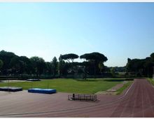 Pista di atletica all’aperto con corsie rosse che curva attorno a un grande prato centrale, con pedane e materassi per salto in alto. Sullo sfondo si vedono alberi alti e un cielo sereno.