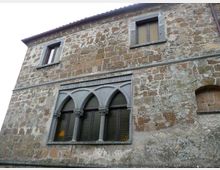 Facciata di un edificio storico in pietra con intonaco consumato, due finestre rettangolari al piano superiore e una grande finestra a tre archi ogivali con colonnine al piano inferiore. Cornici e decorazioni in pietra grigia evidenziano l’architettura.