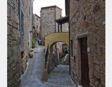 Vicolo stretto in un borgo medievale con edifici in pietra e pavimentazione lastricata, che si biforca tra una rampa e una scalinata. Un arco-ponte intonacato collega due costruzioni sopra il passaggio, con una torre in pietra sullo sfondo.