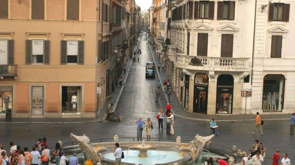 Piazza urbana con una grande fontana ellittica in pietra in primo piano e una lunga strada lastricata che si estende tra palazzi storici a più piani con persiane e vetrine. L’incrocio è ampio e la prospettiva della via conduce verso edifici più lontani, con alcuni veicoli lungo la carreggiata.