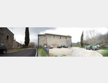 Strada asfaltata con curva accanto a un parcheggio in ghiaia davanti a un edificio in pietra rettangolare con tetto a due falde. Nel piazzale ci sono alcune auto, una recinzione metallica e alti cipressi, con alberi spogli sullo sfondo.