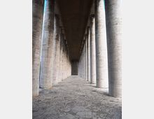 Lungo porticato monumentale con alte colonne cilindriche in pietra disposte in file parallele, che creano una prospettiva verso il fondo. Pavimentazione in lastre di pietra irregolari e soffitto piano in cemento sopra il corridoio.