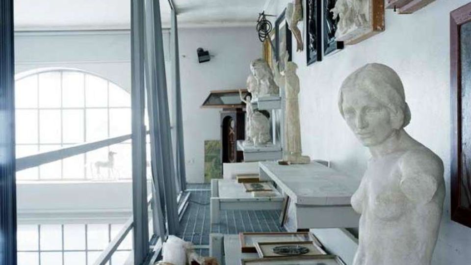 Corridoio in un edificio adibito a deposito o laboratorio museale, con passerella metallica affacciata su grandi finestre ad arco. Lungo la parete sono disposte statue e bassorilievi, con quadri appoggiati su tavoli e alcune scatole sul pavimento.