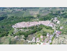 Veduta aerea di un borgo collinare con case in pietra addossate e tetti in coppi, circondato da boschi, campi coltivati e uliveti. Strade tortuose e abitazioni sparse si diramano dal centro abitato nella campagna circostante.