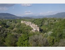 Veduta panoramica di un piccolo borgo con case in pietra addossate su un crinale, circondato da una fitta vegetazione. Sullo sfondo si vedono colline e montagne sotto un cielo parzialmente nuvoloso.