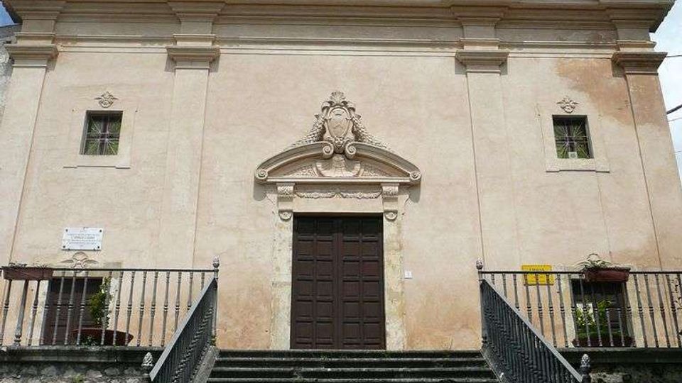 Facciata di una chiesa in stile barocco con intonaco chiaro, portale centrale in legno sormontato da un frontone decorato e finestra superiore, con statue sul coronamento. Una lunga scalinata in pietra con ringhiere metalliche conduce all’ingresso, davanti a un muretto in pietra e sotto un cielo parzialmente nuvoloso.