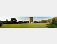 Ampio prato verde in un parco, con alberi lungo i bordi e una torre in mattoni con merlature e scalinata in pietra al centro. Sullo sfondo si vede una fila di alberi e alcuni piccoli edifici bassi sulla destra.
