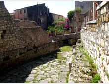 Vicolo stretto in un borgo storico con pavimentazione in pietra irregolare, fiancheggiato da alte mura in pietra e resti murari. Sullo sfondo si vedono edifici antichi e una piccola area verde con vegetazione e fiori vicino a un muretto e a una passerella.