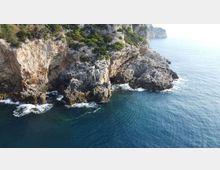 Scogliera costiera rocciosa con pareti di pietra e vegetazione mediterranea, affacciata su mare blu. Onde che si infrangono sugli scogli e promontorio che si estende verso l’acqua.