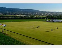 Ampio campo da polo in erba con bandiere su pennoni e strutture con tende e tribune a bordo campo. Sullo sfondo si vedono campi coltivati, una fila di alberi, un centro abitato e colline sotto un cielo sereno.