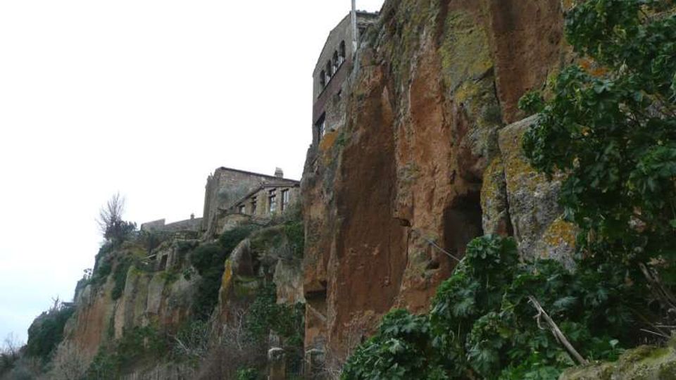 Borgo con edifici in pietra arroccati sul bordo di una alta rupe di tufo rossastro, con pareti rocciose verticali e vegetazione fitta sulle scarpate. In basso si intravede un sentiero con ringhiera ai piedi della scogliera.