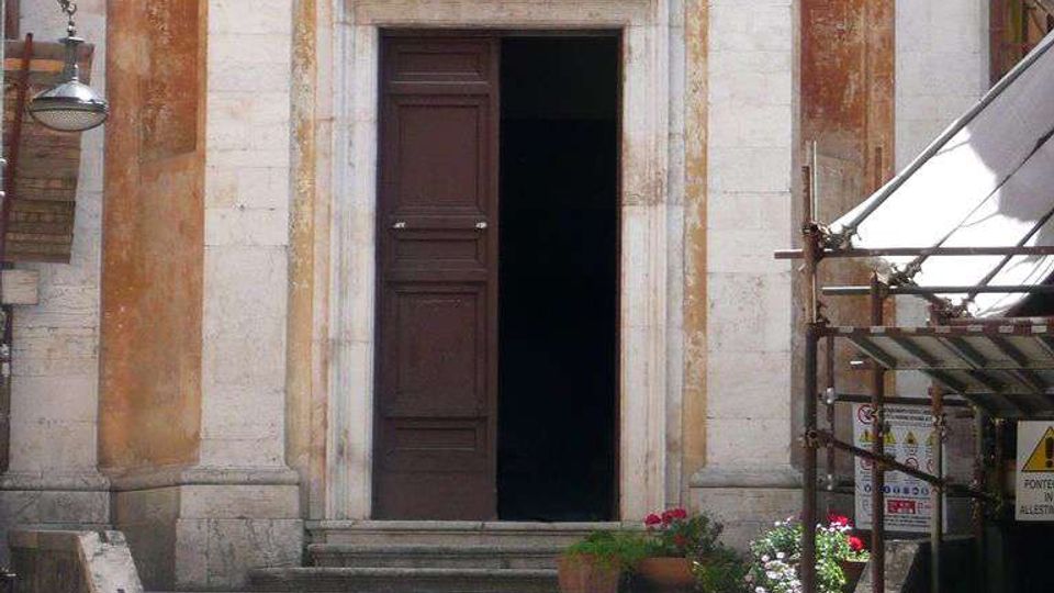Ingresso di un edificio storico con ampia scalinata in pietra che conduce a un portale con cornice in marmo e porta in legno scura parzialmente aperta. La facciata intonacata presenta nicchie verticali, lampioni a muro e alcune luci decorative, con impalcatura e cartelli sul lato destro.