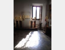 Sala museale con pareti bianche e pavimento in pietra, illuminata dalla luce che entra da una grande finestra ad arco con grate. Diverse teste scultoree su piedistalli e rilievi in pietra sono esposti lungo le pareti e su piattaforme in legno.