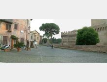 Piazza lastricata in pietra con edifici storici lungo il lato sinistro e una piccola chiesa sullo sfondo. A destra si vedono mura in mattoni con merlature e un grande albero e arbusti che delimitano lo spazio aperto.