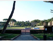 Vista di un impianto sportivo all’aperto con pista di atletica e campo centrale, circondato da siepi e alberi ad alto fusto. Sullo sfondo si vedono antiche mura in pietra e un’area verde con strutture e illuminazione da stadio.